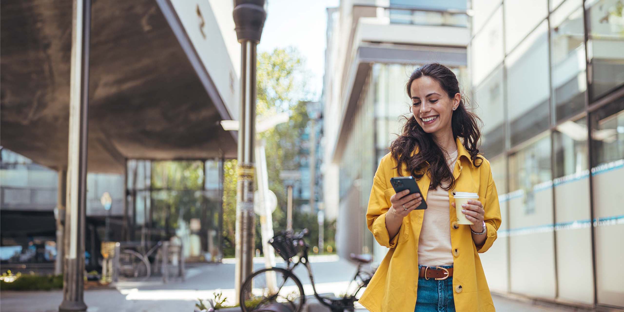 Eine lächelnde Frau in einem gelben Mantel und Jeans geht eine Stadtstraße entlang und hält ein Smartphone und eine Kaffeetasse in der Hand. Im Hintergrund sind moderne Gebäude und ein Fahrradständer mit einem Fahrrad zu sehen. Sie scheint mit ihrem Telefon beschäftigt zu sein.