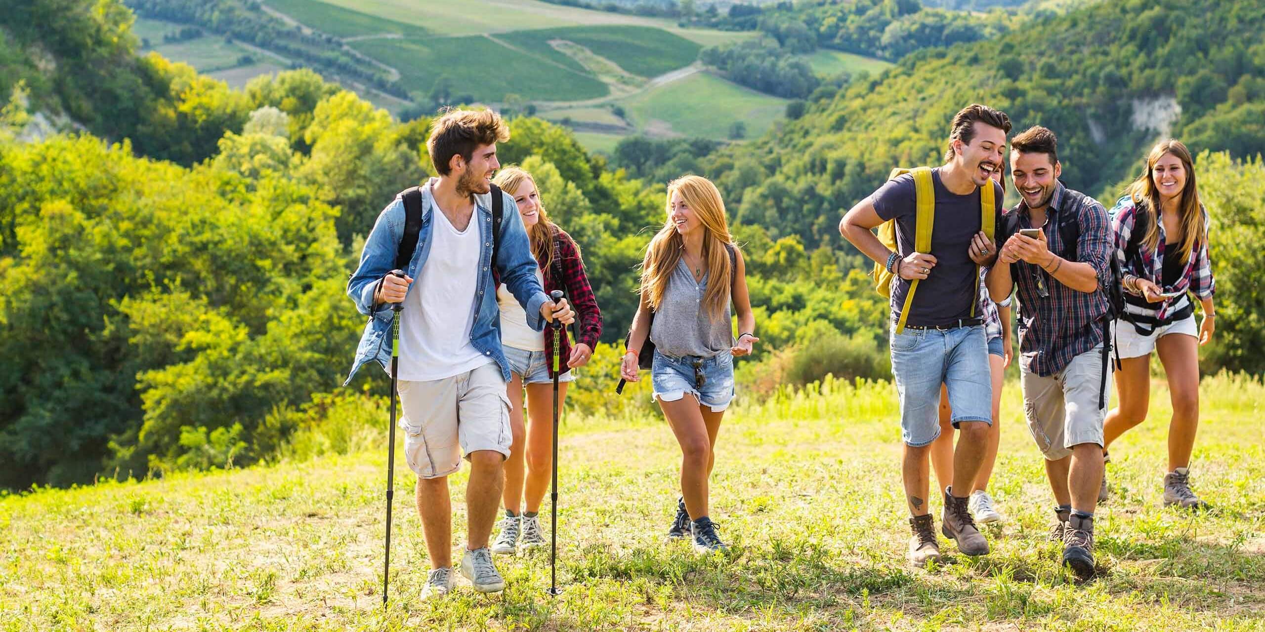Eine Gruppe von sechs Personen, drei Männer und drei Frauen, wandert durch eine malerische, grüne Hügellandschaft. Sie lächeln und lachen, tragen lässige Outdoor-Kleidung, einige tragen Rucksäcke. Der Hintergrund zeigt üppiges Grün und sanfte Hügel.