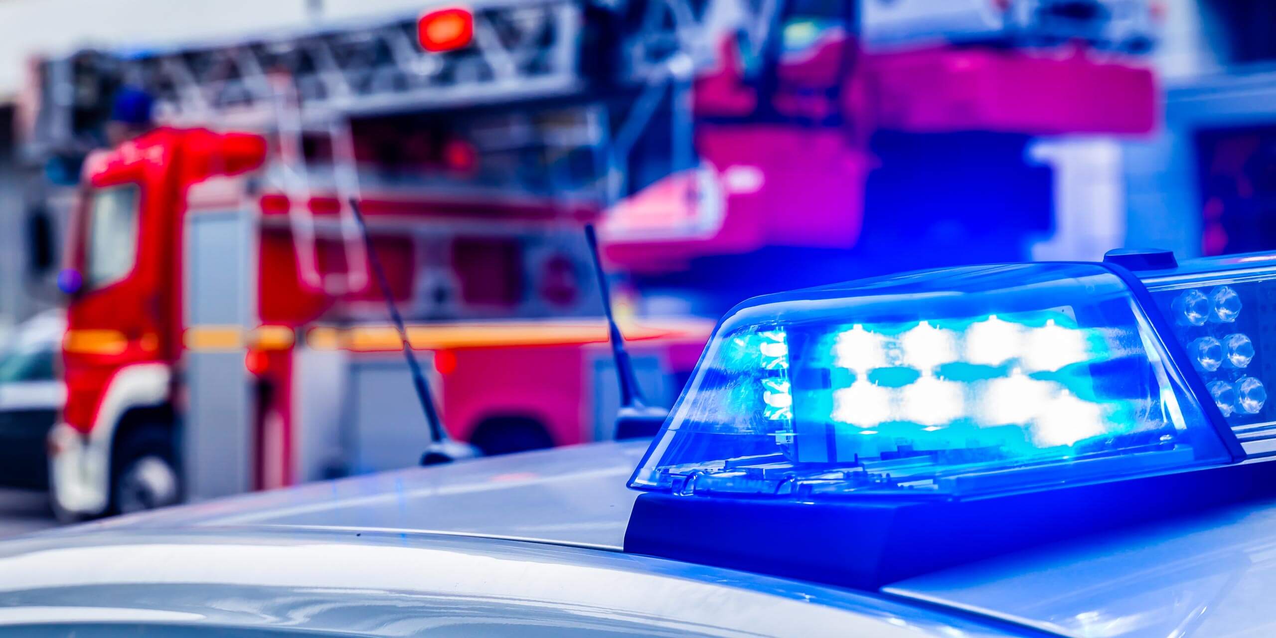 Nahaufnahme der blauen Sirene eines Polizeiautos mit einem verschwommenen roten Feuerwehrauto im Hintergrund. Die Einsatzfahrzeuge stehen auf einer Straße, die wie eine Stadtstraße aussieht, was auf eine aktive Einsatzstelle hinweist.